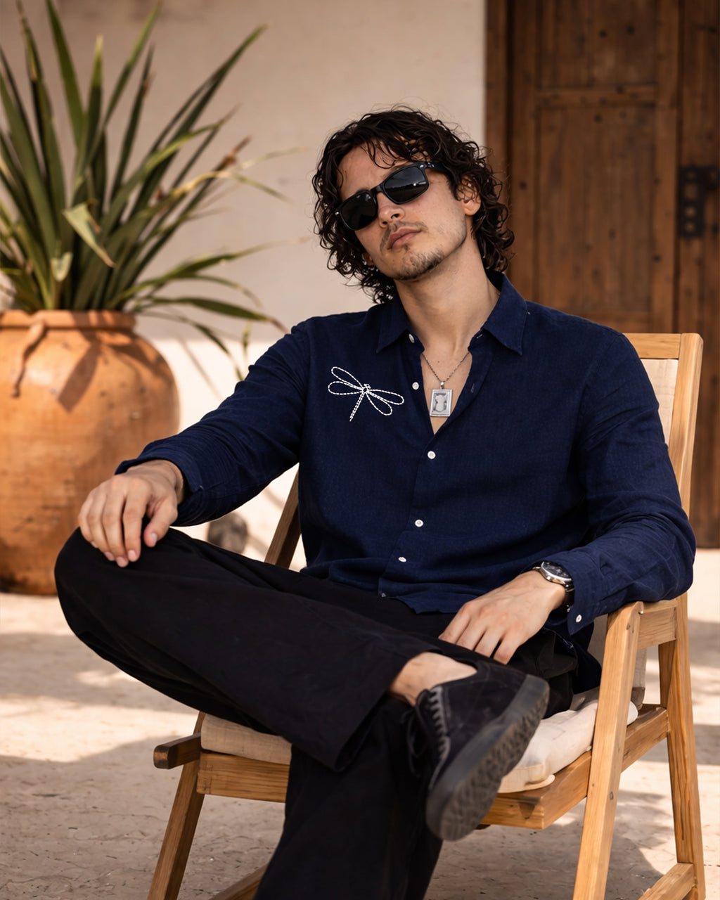 Man sitting on a wooden chair wearing sunglasses and a dark blue shirt with a plant in the background.