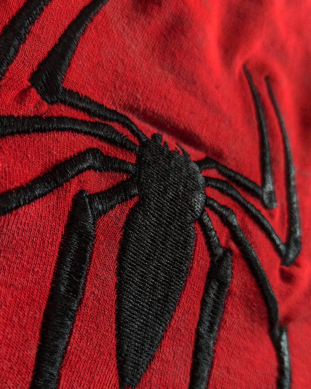 Close-up of a red fabric surface with a large black embroidered spider motif, likely part of a Spider-Man themed garment or accessory.
